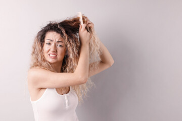 Fototapeta premium Unhappy woman trying to brush her tangled hair on light grey background, space for text