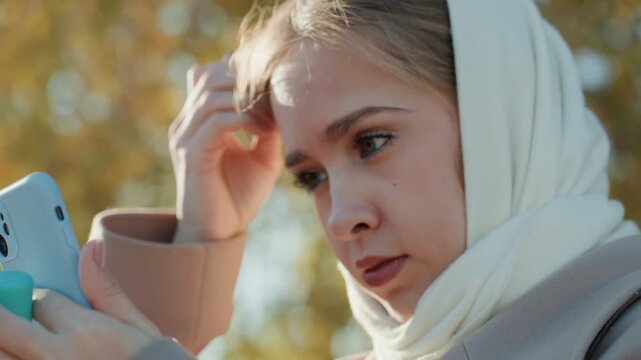 Caucasian woman outdoors checking phone on park bench, scrolling messages, adjusting scarf and hair, applying lipstick between texts, influencer vibe, autumn leaves and golden sunlight, focused
