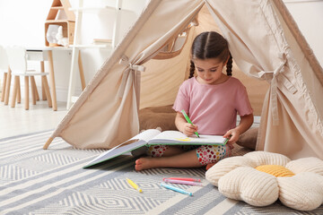 Little girl drawing in sketchbook near toy wigwam at home © New Africa