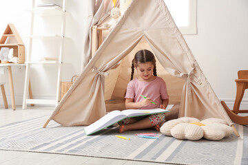 Little girl drawing in sketchbook near toy wigwam at home © New Africa