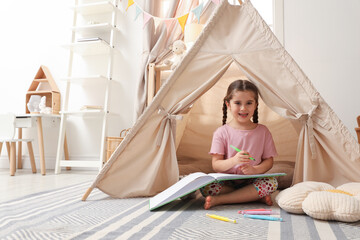 Little girl drawing in sketchbook near toy wigwam at home, space for text © New Africa