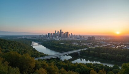 Naklejka premium Cityscape skyline from high hill at sunset. River flows through green forest area. Modern city buildings reflect sunset light. Road bridge crosses water. Clear sky shows soft orange glow.