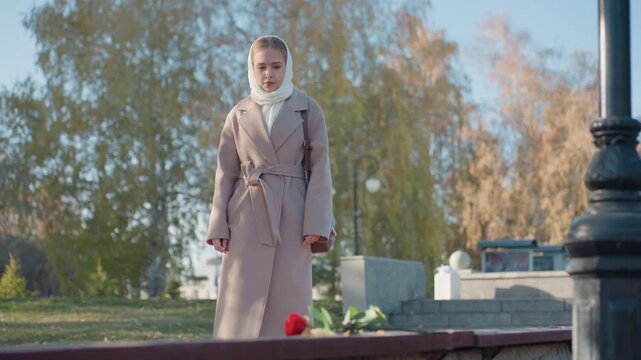 White woman at cemetery placing roses in pale coat and scarf, daughter visiting loved one, walking along stone steps, pausing by headstone under birch trees, kneeling to lay bouquet on grave, touching