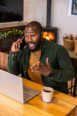 Obraz premium African American man talking on phone, gesturing, working at laptop in home office with coffee mug