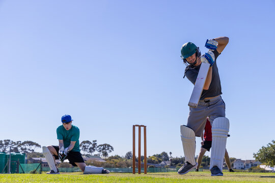 Male batsman wearing green helmet and pads swinging cricket bat on cricket pitch near wooden stumps