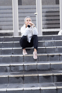 Senior woman sitting on stone steps in urban plaza, wearing hoodie, resting and gazing after rain