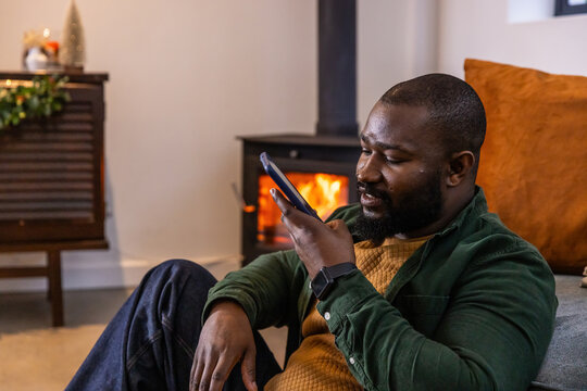 African American man in 30s holding smartphone to face, sitting by wood-burning stove