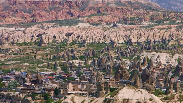 A wide-angle shot of the iconic "fairy chimneys" and ancient cave dwellings in the historic region of Cappadocia, Turkey. The landscape features unique geological formations and a small village nestle