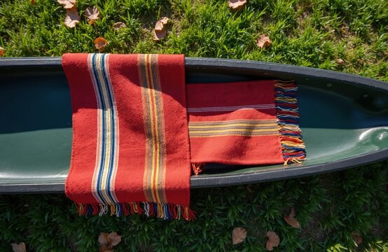 Striped woven sashes rest in a green canoe on grassy ground. Traditional heritage textiles feature bright colorful fringe details. Items evoke canadian voyageur history and adventure.