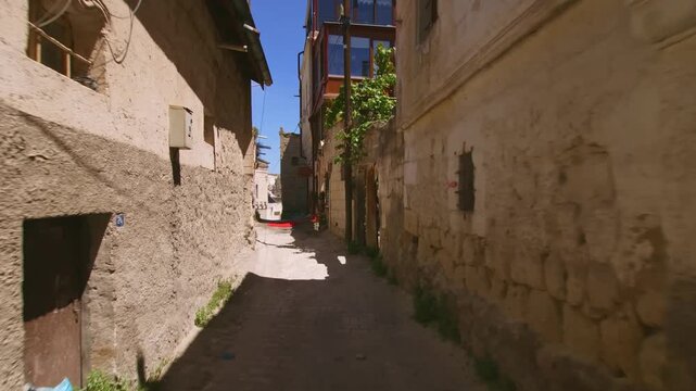 A point-of-view shot moving through a narrow, sunlit alleyway bordered by traditional, aged stone and mud-brick walls in a historic neighborhood of Turkey. The scene captures the authentic, rustic cha