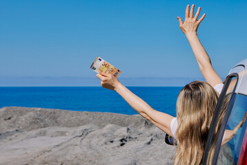 Happy woman leaning out of car window holding 50 euro banknotes and waving at the sea. Concept of travel budget, vacation freedom, and wealth. © beautiful-photo.de