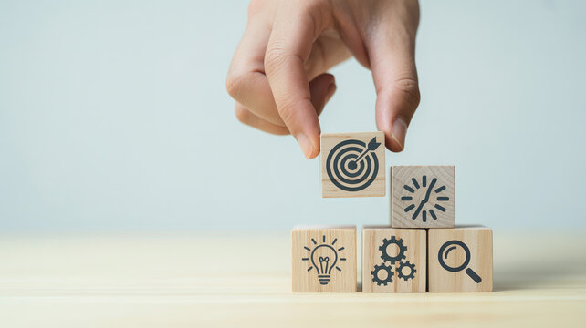 A hand placing a wooden block with a target icon on top of a stack of blocks with various business strategy symbols.
