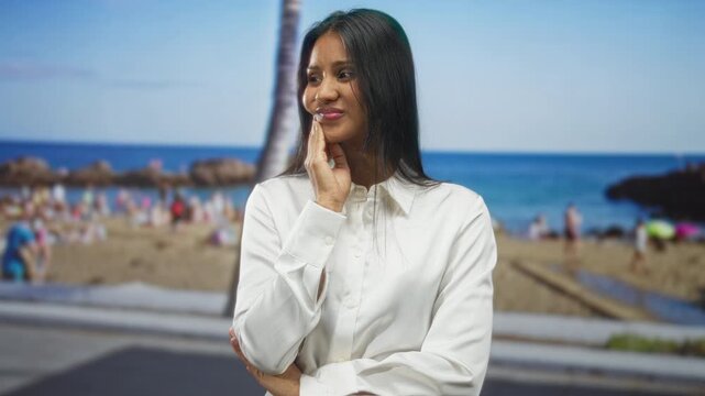 Woman touching cheek with hand, smiling in white shirt at a crowded sandy beach promenade under palm tree; serenity reflection.