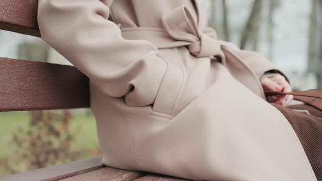 White woman beige coat sitting on bench, commuter checking phone notifications, slipping hand into pocket, retrieving card, scrolling messages, adjusting belt, autumn park backdrop, soft overcast