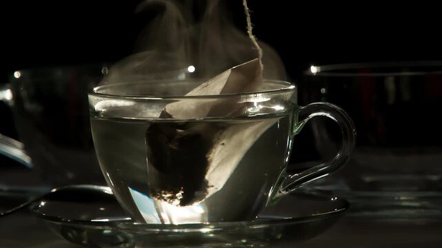 Tea Bag in a Transparent Cup. Teabag falls in a transparent cup with hot water on a dark background.  Water is painted in the color of tea