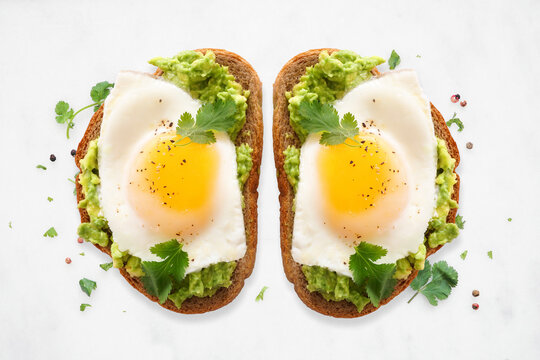 Healthy avocado toast with sunny side up fried eggs on sourdough bread. Top view on a white marble background.