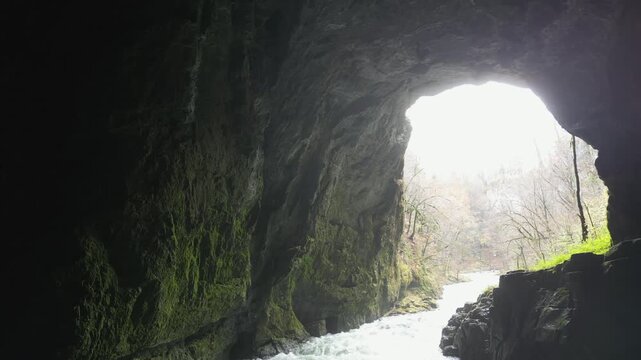 Weaver Cave or Tkalca jama, Slovenia: Stunning view from inside a dark, mossy cave looking out at a powerful river flowing into the daylight. The rocky cavern frames the scene