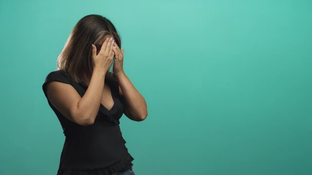 Woman covers her face with hands while wearing glasses, standing against a teal studio wall in a candid, uneasy pose; embarrassment vulnerability.