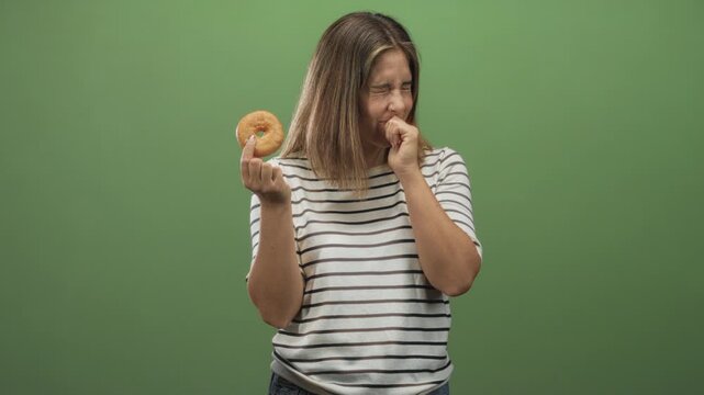 Young hispanic blonde woman holding glazed donut with hand to mouth and gagging against green studio backdrop; disgust regret.
