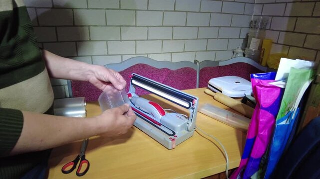 A person sets up a vacuum sealer on a kitchen table. The person prepares to seal food items while surrounded by kitchen tools and appliances in the evening light.