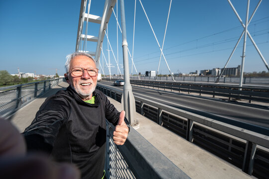A happy senior sport vlogger in sportswear takes a selfie and gives a thumbs up while exercising on a modern urban bridge during his morning run.