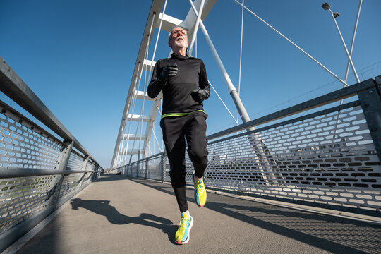 A fit senior man in black athletic wear and neon sneakers is jogging across a white architectural bridge during a morning exercise session.