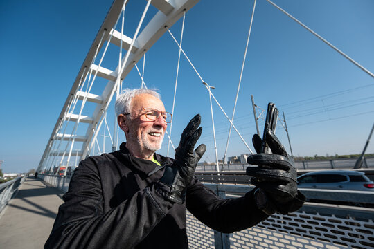 Side view of happy senior sport vlogger in sportswear takes a selfie and gives a thumbs up while exercising on a modern urban bridge during his morning run.