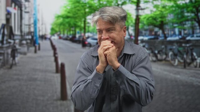 Man clasping hands to mouth on a street, biting knuckles with tense posture and visible hands near face; anxiety vulnerability.