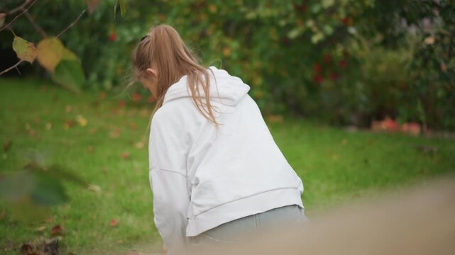 White girl hoodie bending over grass, quiet backyard garden scene collecting fallen leaves, ponytail braid, soft autumn light, contemplative mood, closeup framing, natural textures for stock footage