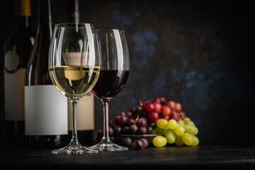 Fototapeta premium A dark, elegant still life of White and Red wine in a glass with wine bottles and fresh grapes in the background. Perfect for wine-themed compositions