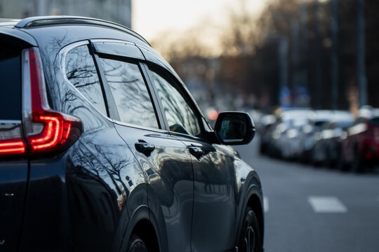 Dark SUV seen from the rear side with glowing taillight, side mirror and reflective windows on a city street lined with parked cars and soft evening blur
