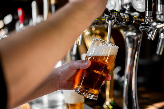 A bartender's hand pouring a fresh, amber-colored lager beer from a polished chrome tap into a glass with a frothy white head in a dimly lit bar