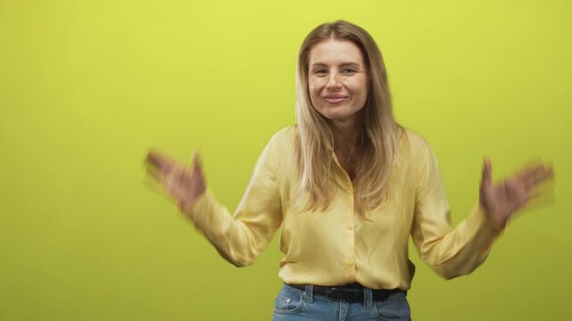Blonde woman sticks tongue out and lifts hands to her head in an antler gesture, wearing yellow shirt and jeans against a lime studio backdrop; playful mischief laughter.