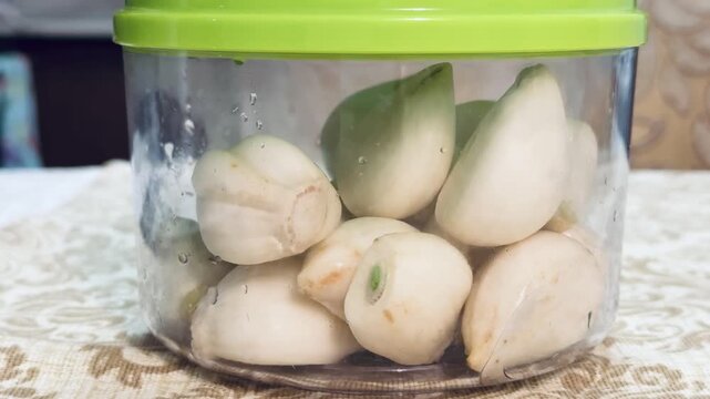 Vacuum container peeled garlic on tablecloth, green lid, clear plastic jar, patterned beige tablecloth, blurred kitchen.