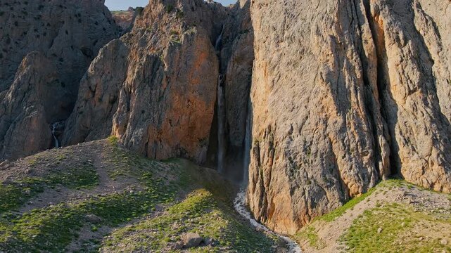 Sunlight glints off rugged canyon walls. Vines cling to rock, hinting at life's resilience. Narrow trail winds through the terrain. The scene feels untouched, wild, and ancient
