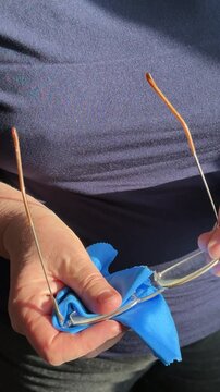 Closeup woman cleaning eyeglasses with blue microfiber cloth, sunlight catching gold frames and hands, slow deliberate wiping.