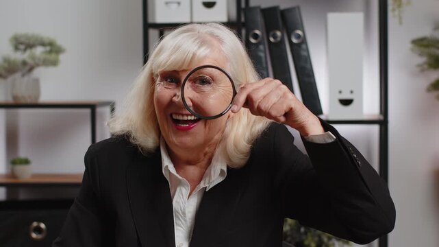Investigator researcher businesswoman looking through magnifying glass with wow expression with big zoomed funny eye, searching, analyzing. Professional Caucasian freelancer grandma looking at camera.