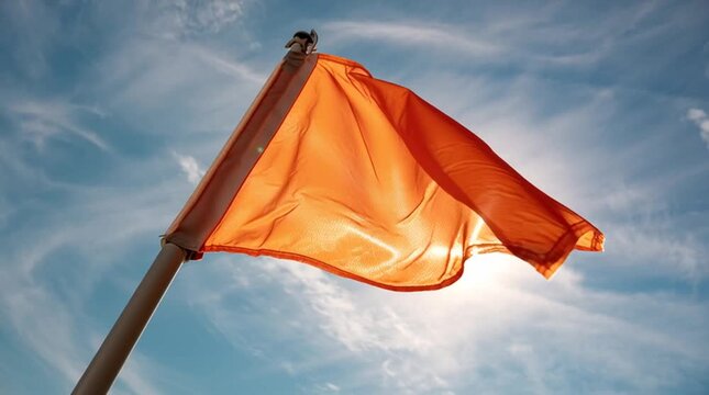 A vibrant orange flag waves in the wind against a bright blue sky with wispy clouds in this dynamic video.