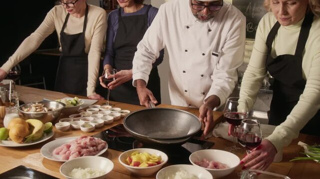 Biracial chef in white uniform setting up pan on stove during cooking master class with group of middle aged women in restaurant