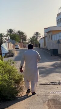 Rear view of a man in traditional white attire strolling down a bright, paved street in a Middle Eastern or South Asian town, framed by lush palm trees, green bushes, and local architecture.