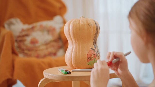 Caucasian girl painting floral pumpkin design bright paints and steady brushwork closeup of hand adding petals warm sofa and cushion backdrop playful harvest craft scene child artist exploring color