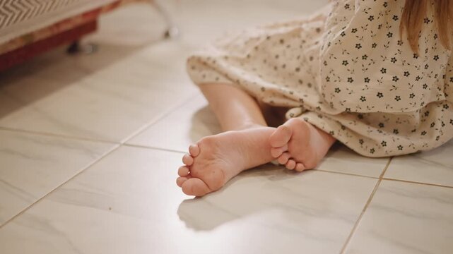 Bare feet child sitting on tile, soft sunlight across patterned dress, relaxed posture near radiator, closeup of toes and floor texture, warm cozy domestic mood suggesting quiet daydream