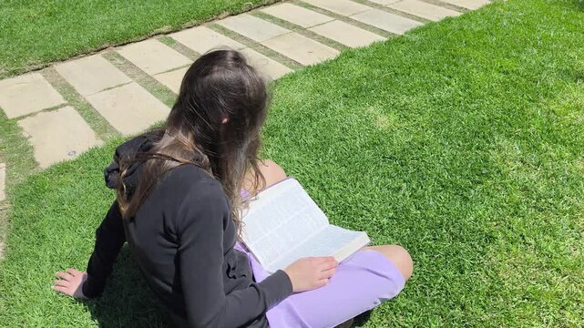 Young Christian Teen Girl Sitting Reading Bible in a Green Park with Grass - Slow Motion Half Speed
