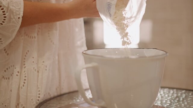 Caucasian woman pours flour into white ceramic bowl, stream of flour cascades from bag into bowl on scale, sunlight backlight emphasizes texture and motion, lace dress sleeve, focused measuring