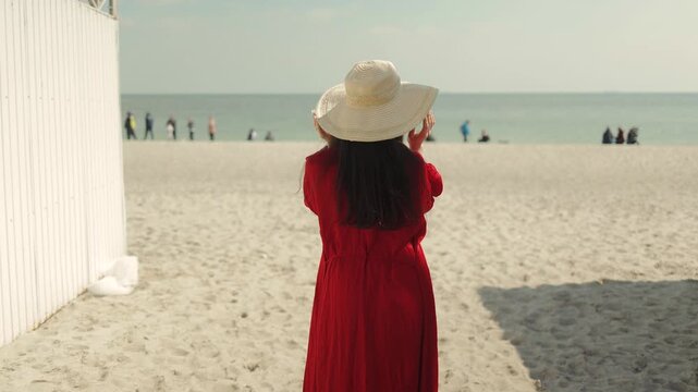 A beautiful woman in a red beach robe and straw hat on the sandy shore. Summer, vacation, ocean, lifestyle, travel, fashion, resort, elegance, sunny, city beach, heat, tourism, sea