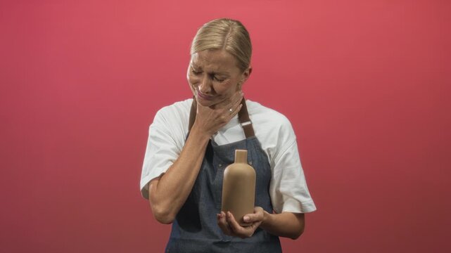 Middle aged blonde pottery artisan wearing denim apron holds unglazed clay bottle with both hands and touches neck in studio; discomfort.
