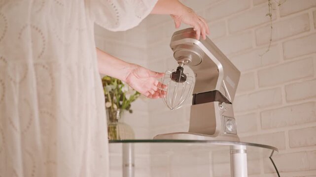 Hands attach beater to mixer on glass table by white brick wall, vase with flowers and tray of baked goods visible, warm soft light, home baker preparing pie crust and ingredients with calm ritual