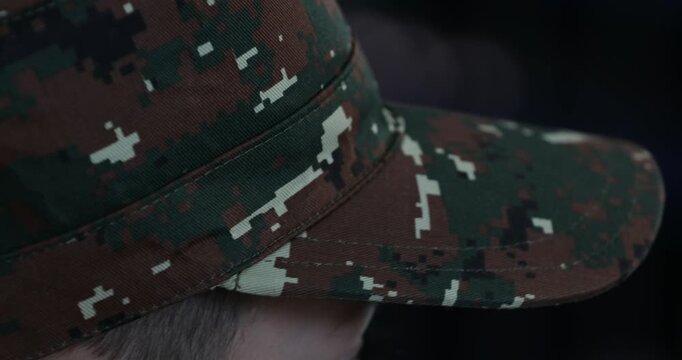 A close-up shot of a female hand gently adjusting the brim of a military camouflage cap. The pixelated camo pattern is in sharp focus against a dark background.