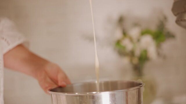 White home baker mixing pumpkin pie filling in stainless bowl, gentle hand stirring with whisk and scraping batter, smooth custard dripping back into bowl, soft sunlit kitchen with blurred flowers,