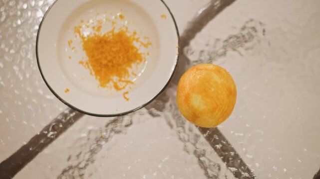 Overhead shot of orange and zest on enamel plate, top view composition with glass table reflections, clean minimal styling, interplay of whole fruit and grated peel for garnish, calm natural light